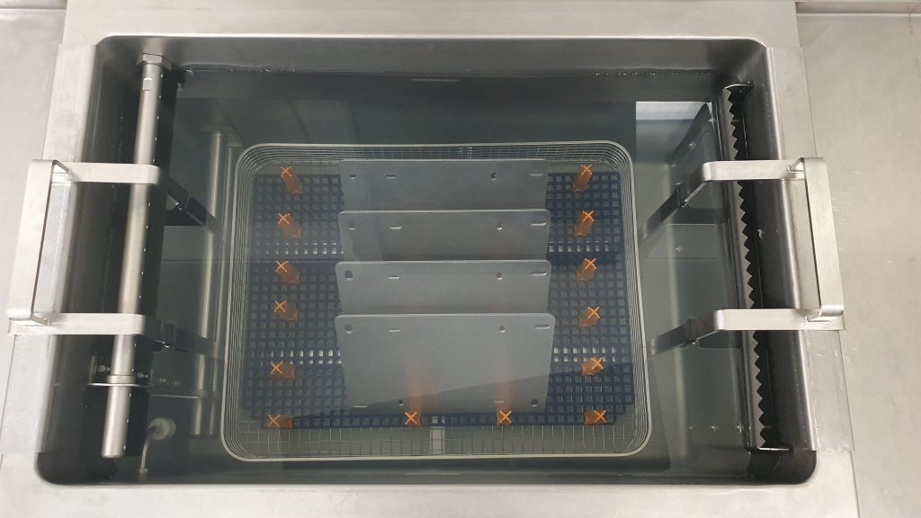 Vier Metallplatten aus einem Blechfertigungsprozess, die aufrecht in einem Drahtkorb stehen, der in ein Ultraschallreinigungsbecken eingetaucht ist.